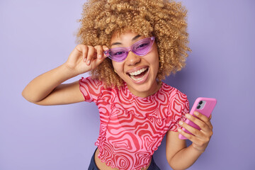 Wall Mural - Positive joyful woman with curly bushy hair wears trendy sunglasses and t shirt uses mobile phone chats in app laughs happily isolated over purple background. People emotions and technologies