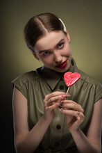 Portrait Of A Young Attractive Girl With A Smooth Hairstyle And Hairpins On Her Blonde Hair In A Green Stylish Dress With A Red Sweet Candy Lollipop In The Shape Of A Heart In Her Hands. Close-up