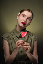 Portrait Of A Young Attractive Girl With A Smooth Hairstyle And Hairpins On Her Blonde Hair In A Green Stylish Dress With A Red Sweet Candy Lollipop In The Shape Of A Heart In Her Hands. Close-up