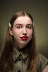 Wall Mural - Portrait of a young attractive girl with a smooth hairstyle and clips on her blonde hair in a stylish green dress with expressive red lips. Close-up, green background