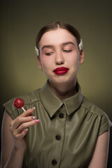 Wall Mural - Portrait of a young attractive girl with a smooth hairstyle and clips on her blonde hair in a green stylish dress with a red sweet candy lollipop in her hands. Close-up, green background