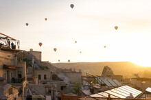 Sunrise At Goreme Town Of Cappadocia, People Watching Fly Of Hot Air Balloons On Roofs, Fairy Chimneys And Houses Around. Nevsehir, Turkey. 