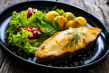 Fish dish - fried halibut with fried potatoes, cherry tomatoes and leafy greens on wooden table
