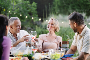 Mature friends having dinner around table in a garden on a summer evening, drinking wine, having fun talking and laughing