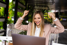 Young Woman Rejoices In Winning Having Raised Her Hands Up Sitting In Front Of Laptop In Cafe