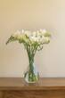 Vertical closeup of white freesia flowers in glass vase on oak table against beige wall (selective focus)