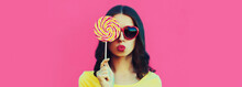 Portrait Of Happy Young Woman Covering Her Eyes With Colorful Lollipop Wearing Heart Shaped Sunglasses Blowing Her Lips On Pink Background, Blank Copy Space For Advertising Text
