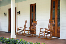 Three Vintage Brown Wooden Rocking Chairs Are On The Veranda Of An Old White Colored Country House With A Wrap Around Porch. There's A Flower Bed And Garden In Front Of The Building With Green Grass. 
