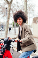Smiling Young Woman Rides An Electric Bike From A Rental Station, Active Lifestyle And Sustainable Mobility Concept