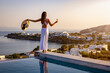 A beautiful woman in white dress at the pool enjoys the sunset behind the mediterranean sea with a glass of wine