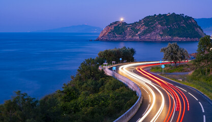 Wall Mural - Car lights on the road on the coast of Euskadi. Getaria lighthouse at dusk.