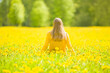 Young adult blonde woman sitting alone on grass at meadow of fresh yellow blooming dandelions in warm sunny spring day. Thinking about life in restful moment. Peaceful atmosphere in nature. Back view.