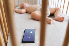 Selective Focus Of Little Boy In Crib Falling Asleep With The Help Of Application For Baby Sleep On Phone Lying Next To Him While Mother Cooking Dinner At Kitchen. Childcare And Technology