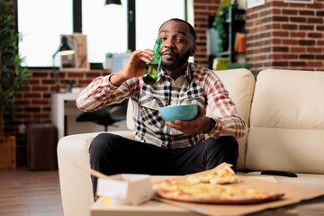 Wall Mural - Happy guy drinking alcoholic beer from bottle and holding bowl of chips, watching film on television program. Enjoying beverage with takeaway delivery food and snacks, entertainment.