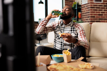 Wall Mural - Young adult drinking alcoholic beer from bottle and eating takeaway meal from fast food delivery package, watching movie on tv. Enjoying drink and takeout meal in front of television.