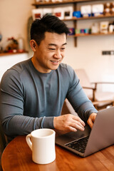 Happy adult asian man smiling and using laptop in cafe indoors