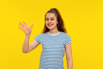 Wall Mural - Friendly cheerful brunette girl smiling, saying hi and waving hand to greet person, make hello gesture, welcome someone, standing in white-blue striped t shirt over yellow background