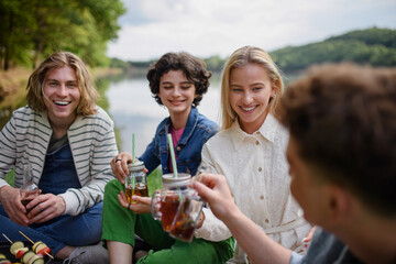 Group of young friends having fun on picnic near a lake, sitting on blanket and toasting with drinks.