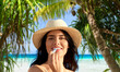 beauty, cosmetics and summer holidays concept - portrait of smiling young woman in bikini swimsuit and straw hat applying lip balm over tropical beach and palm trees background in french polynesia