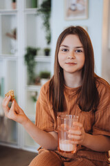 Brunette caucasian teenage girl at home in light brown pajamas holding cookie and glass of milk looking at camera against blurry living room. Teenage on breakfast time before school. Girl portrait.