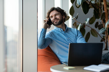 Poster - Handsome mature businessman in formal wear using laptop and speaking on smartphone at modern office, free space
