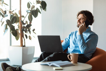 Poster - Smiling mature businessman talking on cellphone, using laptop, sitting in bean bag chair at company office, copy space