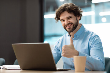 Poster - Portrait of happy businessman sitting at desk, using laptop computer and showing thumb up gesture, working at office