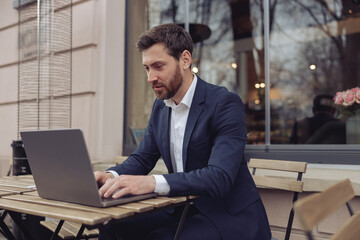 Wall Mural - Caucasian handsome man working at laptop outdoors in cafe. Texting on keyboard. Business concept.