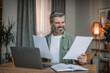 Smiling caucasian senior man with beard works on laptop with documents in living room interior