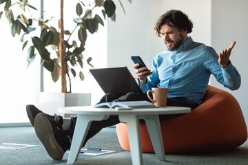 Wall Mural - Annoyed mature businessman with laptop looking at cellphone screen and gesturing angrily, working at office, copy space