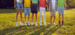 canvas print picture Little kids having fun in a sunny park. Group of unrecognizable boys and girls in summer clothes jumping on green grass all together. Cropped shot, children's legs. Banner or header background