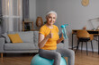 Stay hydrated. Happy senior female holding bottle with fresh water and showing thumb up gesture, sitting on fitball