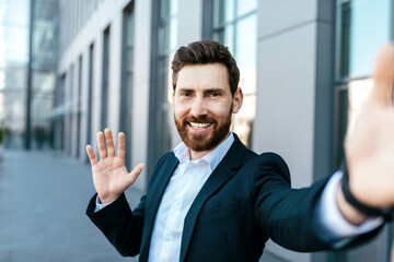 Happy confident handsome european millennial bearded businessman takes selfie, waving hand on street