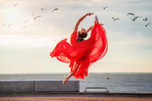 Jumping Ballerina In A Red Flying Skirt And Leotard On Ocean Embankment Or Sea Beach Surrounded By Seagulls In Sky.