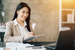 © Kritdanai - Portrait of attractive woman sitting at office desk, writing on notebook and looking at laptop screen