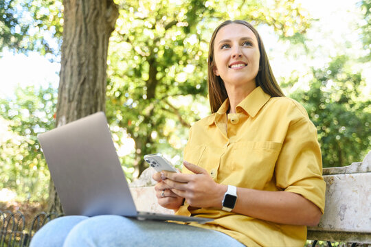 Positive female student wearing yellow casual shirt texting on the smartphone and using laptop computer sitting on the bench, young freelancer woman chatting in social networks