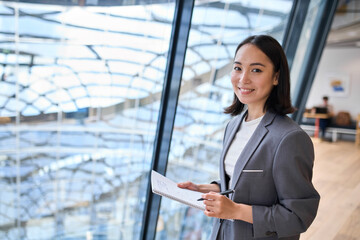 Smiling young Asian business woman manager wearing suit holding notebook standing in modern glass office. Professional executive manager, corporate leader looking at camera. Portrait