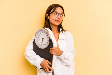 Wall Mural - Young nutritionist woman holding a weighing machine isolated on yellow background looks aside smiling, cheerful and pleasant.