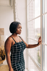 Wall Mural - Young inspired woman standing looking through the window, watching views of city landscape and scenery. People at home. Peaceful morning. Portrait of confident lady girl