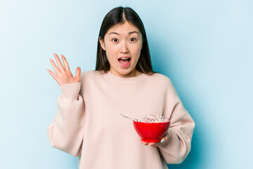 Wall Mural - Young asian woman holding a bowl of cereals isolated on blue background receiving a pleasant surprise, excited and raising hands.