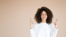 May Dreams Come True. Positive African American Woman With Cury Haircut, Crosses Fingers, Dressed In White T Shirt, Has Amazed Expression, Isolated Over Beige Background, Copy Space For Your Promotion