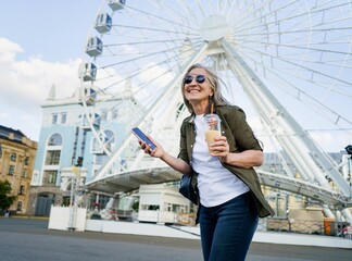 European charming grey haired woman happy enjoying free time after work or traveling holding phone while having juice on the go using plastic cup in city and ferris wheel on background