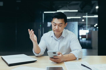 Upset asian businessman working in office, reading bad news on phone, man depressed at work