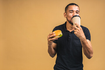 Hungry young african american black man eating hamburger isolated over beige background. Diet or Fast food concept. Drinking coffee.