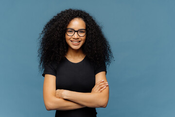 Wall Mural - Waist up shot of smiling Afro American woman has arms folded, wears spectacles and casual black t shirt, enjoys nice conversation with interlocutor, poses over blue studio wall with blank space