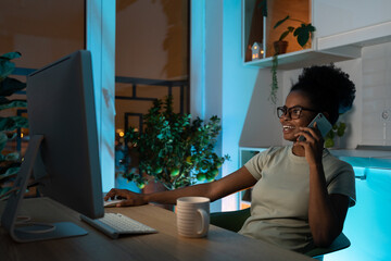 Positive african american woman in glasses using computer mouse, talking on smartphone, working on desktop pc late at night at home office during the night. Remote work concept