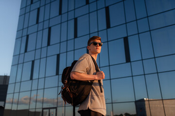 Wall Mural - Portrait of handsome tourist hipster man in sunglasses with bag handy, standing guy near the building in downtown. Calm relaxing moment, summer vacation concept