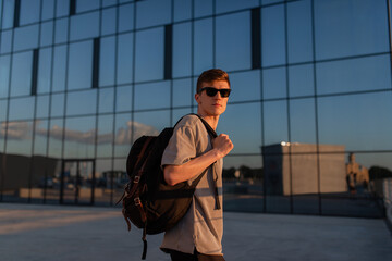 Wall Mural - Portrait of handsome tourist hipster man in sunglasses with bag handy, standing guy near the building in downtown. Calm relaxing moment, summer vacation concept