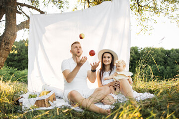 Wall Mural - Happy young family having picnic sitting on white blanket outdoors. Charming mother holding her little daughter in arms while father playing by throwing apples. Family concept.