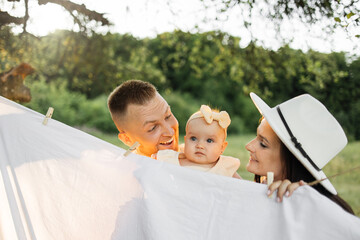 Wall Mural - Beautiful caucasian man and woman standing and embrace with their cute little daughter look at each other through the curtain outdoor. Family time on fresh air.